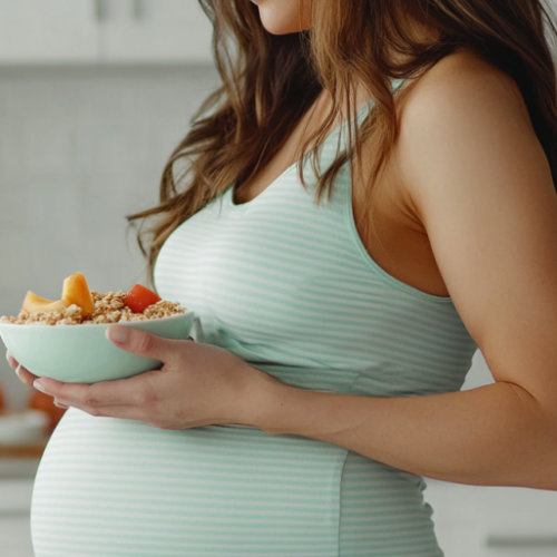 Kobieta o długich brązowych włosach, w topie w biało-błękitne paski, dbająca o dietę w zaawansowanej ciąży, stoi w kuchni z miską owsianki z owocami.